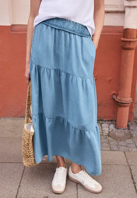 Frau in weißem Top, langer blauer Stufenrock, weiße Sneaker und Korbtasche vor Hauswand.